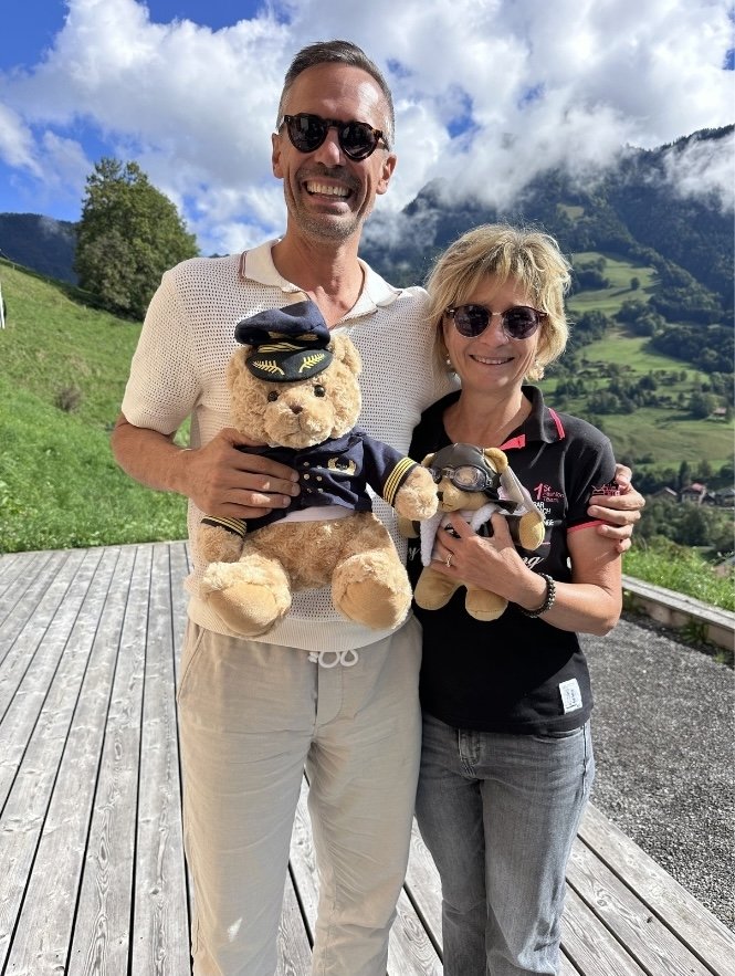 Deux personnes souriantes tenant chacune une peluche d’aviateur, en extérieur, devant un paysage de montagnes.